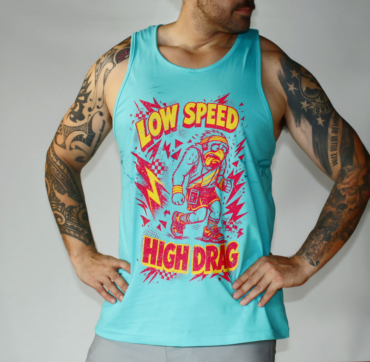 A man wearing a turquoise tank top with a colorful graphic print that reads 'LOW SPEED HIGH DRAG' and shows tattooed arms against a plain gray studio background.