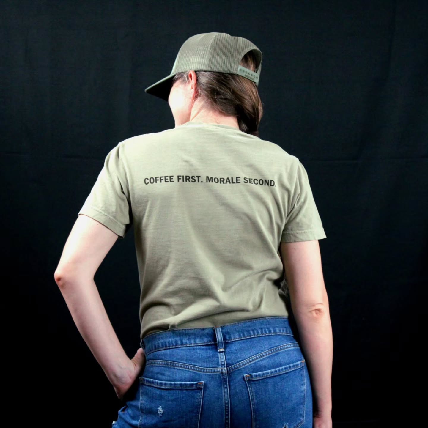 A person shown from the back wearing an khaki t-shirt with the text 'COFFEE FIRST, MORALE SECOND.' printed in black across the upper back, paired with blue jeans and a matching olive green cap against a black background.