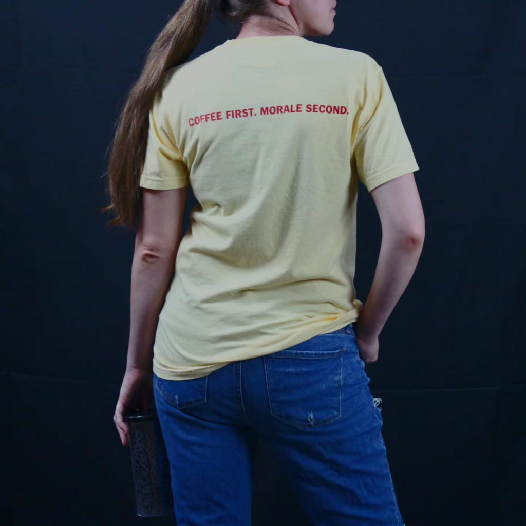 A person shown from the back wearing a pale yellow short-sleeve T-shirt with red text reading 'COFFEE FIRST. MORALE SECOND.